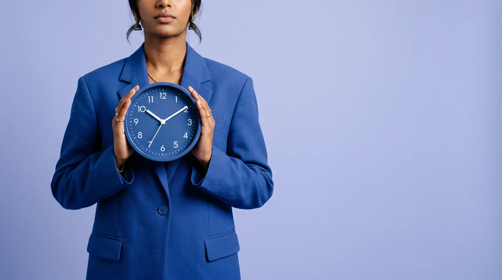 A South African Indian woman in a cobalt blue suit, holding a matte cobalt blue wall clock against her chest in both hands