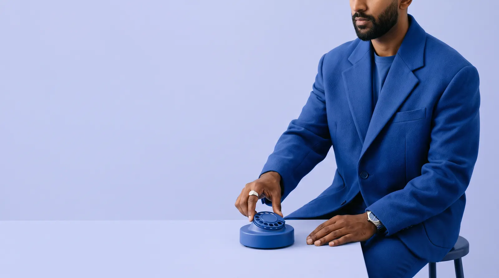 A South African Indian man in a cobalt blue suit, seated at a table, one hand on the dial of a matte cobalt blue rotary telephone